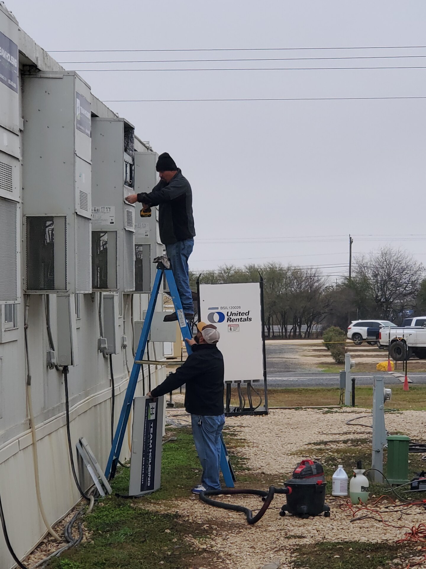 Two Fresh Air technicians on ladders servicing a row of commercial HVAC units with vacuum equipment staged below
